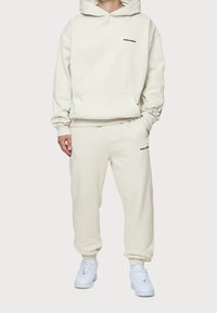 Felpa leggera beige con tasca a marsupio e pantaloni della tuta abbinati indossati da una persona. Stanno in piedi con le mani nelle tasche, indossando sneakers bianche.
