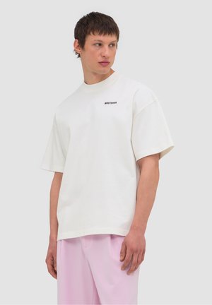 Young man wearing a loose white T-shirt with small black text on chest and light purple pants, looking to his side against plain background.