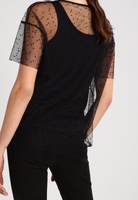 Mujer vistiendo una blusa sin mangas negra con una sobrecamisa corta y transparente que presenta un pequeño estampado de lunares, vista desde la espalda.