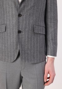 Blazer grigio a righe sottili realizzato in lana texturizzata, con due bottoni, colletto a punta e tasche frontali. Indossato sopra pantaloni grigi chiari.