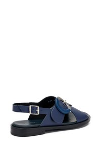 Sandalo in pelle blu con punta chiusa, lati aperti, cinturino alla caviglia regolabile e dettaglio circolare. Suola piatta con finitura testurizzata.