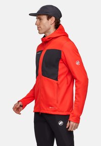 Rød softshell-jakke med sorte detaljer, lynlås-lommer og hætte, med et logo på venstre ærme. Strækbar, tætsiddende design.
