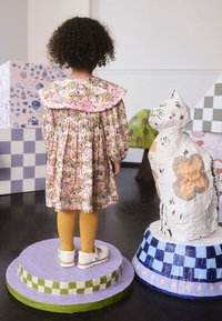 Dieťa oblečené v kvetovaných šatách s veľkým ružovým volánovým golierom, žltými pančuchami a bielymi topánkami, stojace vedľa maľovanej sochy mačky.