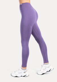 Mujer con leggings morados de cintura alta y zapatillas Nike blancas, de pie con una pierna ligeramente doblada contra un fondo claro y liso.