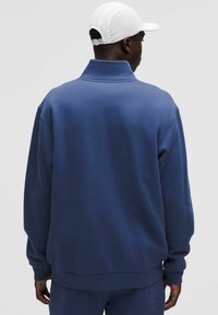 Uomo che indossa un maglione navy a vestibilità comoda e pantaloni abbinati, visto di spalle, con un cappellino da baseball bianco su uno sfondo semplice.
