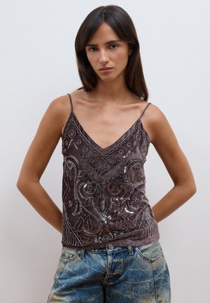 Donna con lunghi capelli scuri che indossa un top scuro senza maniche con paillettes e jeans blu sbiaditi, in piedi davanti a uno sfondo chiaro e uniforme.