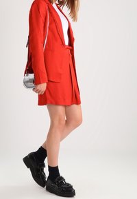 Röd blazer med spetsdetaljer, röd kjol med skärp, kombinerad med svarta klumpiga skor och strumpor. En liten silverfärgad crossover-väska ger extra detalj.