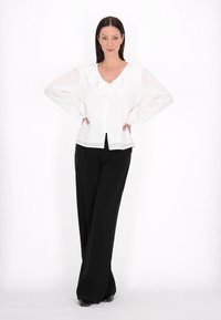 Blusa bianca con scollo arricciato e abbottonatura frontale, abbinata a pantaloni neri a gamba larga. Tessuto liscio e vestibilità sartoriale.