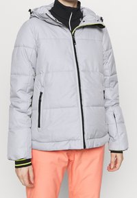 Personne portant une doudoune gris clair avec des détails de fermeture éclair noirs et un pantalon corail, debout devant un fond blanc uni.