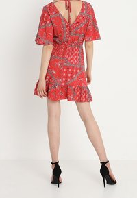 Rotes kurzes Kleid mit Paisleymuster, ausgestelltärmel und tailliertem Schnitt. Mit V-Ausschnitt und gerafftem Saum. Getragen mit schwarzen High Heels.