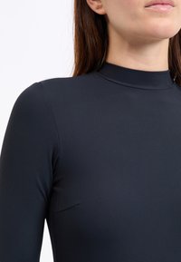 Schwarzes, geripptes Langarmoberteil mit hohem Ausschnitt. Glatte Textur und figurbetontes Design mit dezenten Nahtdetails nahe der Schulter.