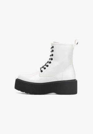 Biały lakierowany botek wojskowy z czarnymi sznurowadłami i masywną czarną podeszwą na platformie. Posiada pętlę na pięcie ułatwiającą zakładanie.