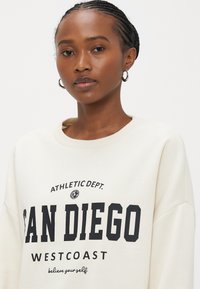 Cremefarbener Sweatshirt mit rundem Ausschnitt und überschnittenen Schultern. Schwarzer Aufdruck mit dem Text "SAN DIEGO WESTCOAST" und grafischen Akzenten.