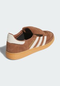 Hnedé tenisky Adidas Spezial s bielymi pruhmi, gumenou podrážkou, semišovým pätou a čiastočne otvoreným jazykom, z pohľadu zo zadnej ľavej strany.