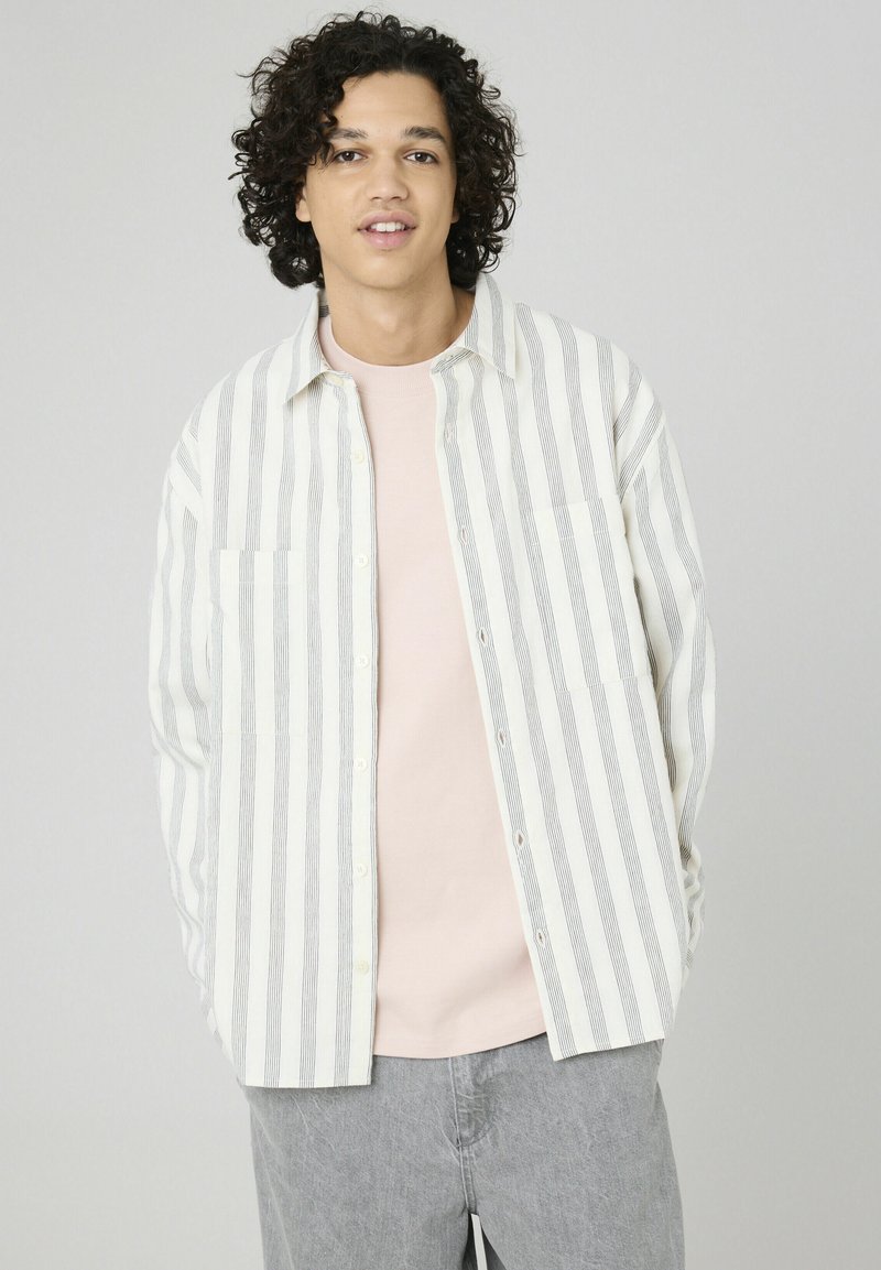 Chemise à rayures blanche et grise en tissu coton, avec deux poches avant, associée à un t-shirt rose et un pantalon gris.