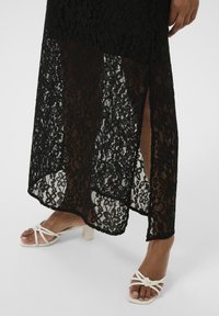 Robe en dentelle noire avec une fente haute et un motif floral, associée à des sandales blanches à talons carrés et fines lanières. Surface plate et texturée.