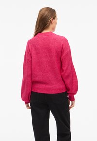 Jasnoróżowy, dziergany sweter o luźnym kroju, z prążkowaną teksturą i obszernymi rękawami. W zestawieniu z czarnymi spodniami z tylnymi kieszeniami.