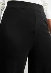 Pantalons noirs à texture lisse, avec une taille haute, des plis à l'avant et une coupe ajustée, vus d'un angle bas.