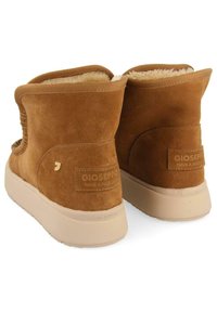 Botas de tobillo de ante marrón con un forro suave, que presentan una suela de goma beige y un detalle del logotipo en la parte trasera. Diseño redondeado con lados elásticos.