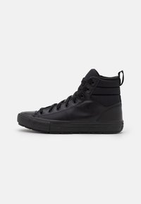 Hoge zwarte sneaker met veterbinding aan de voorkant, gewatteerde enkel, rubberen zool en een aantreklip aan de hak op een witte achtergrond.