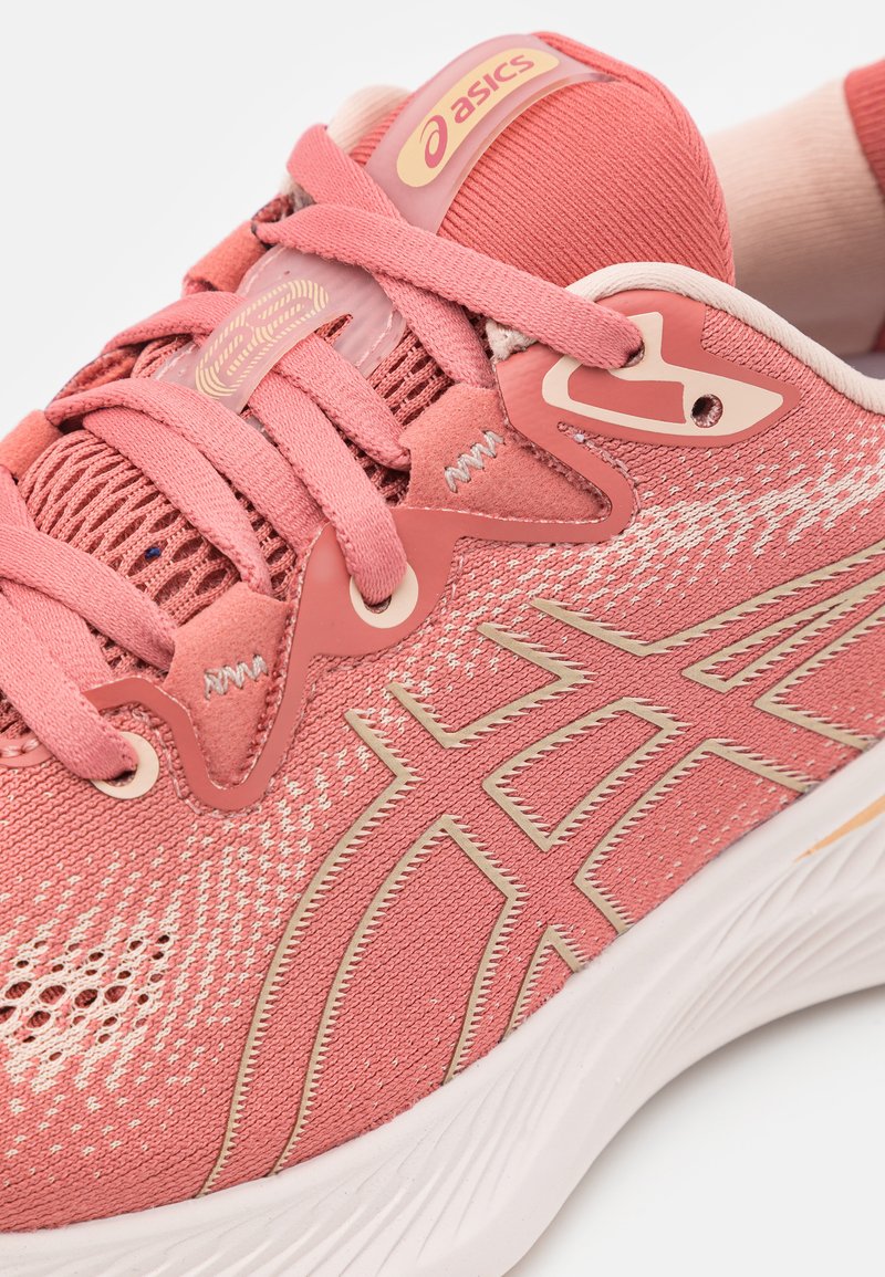 Rosa Sportschuh mit Mesh-Obermaterial, dekorativen Nähten und beigen Akzenten. Verfügt über eine gepolsterte Sohle und ein auffälliges Logo an der Zunge.