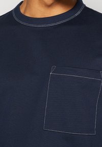 Granatowy t-shirt z krótkim rękawem i okrągłym dekoltem, z naszywaną kieszonką i kontrastującym białym przeszyciem dla dodatkowego detalu i faktury.