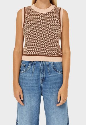 Persoon draagt een mouwloze gebreide top met een geometrisch bruin en beige patroon, gecombineerd met een hoge taille blauwe spijkerbroek, tegen een effen achtergrond.