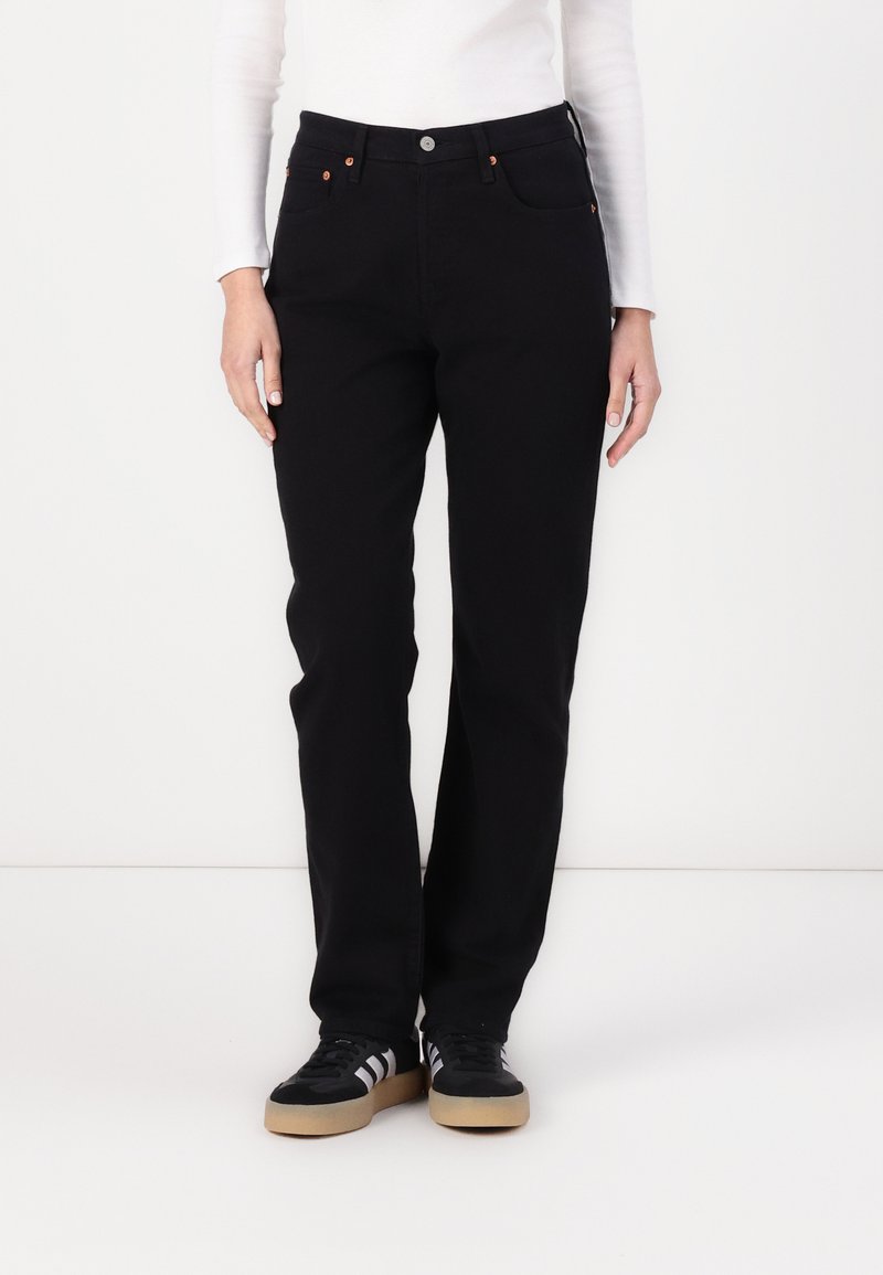 Pantalones de mezclilla negra con un diseño de pierna recta, que presentan detalles de botones de cobre, bolsillos laterales y una cinturilla clásica. Combinados con zapatillas negras.