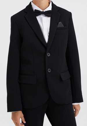 Zwarte blazer met textuur, voorzien van ingesneden revers, twee knopen en zijzakken. Draagt over een wit overhemd, aangevuld met een zwarte vlinderdas.