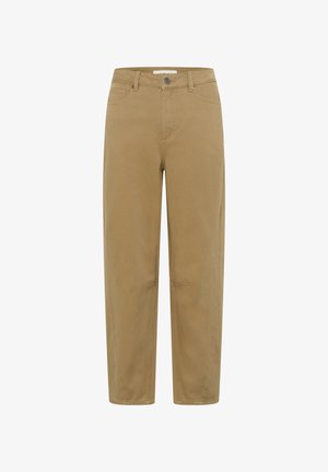 Femme marchant en intérieur portant une veste beige à clous et un pantalon large assorti avec des chaussures noires à bout pointu devant un fond de cadre en bois.