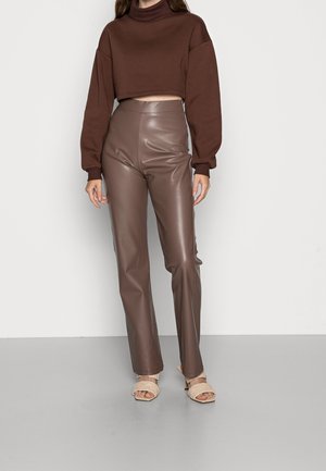 Donna che indossa un maglione marrone a collo alto corto, pantaloni di pelle marrone a vita alta e sandali beige aperti con tacco, in piedi su pavimento grigio.
