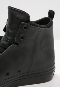 Schwarzer High-Top-Sneaker aus mattem Leder, mit runden Metallösen, strukturierten Akzenten und einem kreisförmigen Logo an der Außenseite.