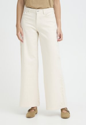 Pantaloni beige a gamba larga con chiusura con bottone e cerniera indossati con ballerine Mary Jane in camoscio marrone su una persona in piedi.