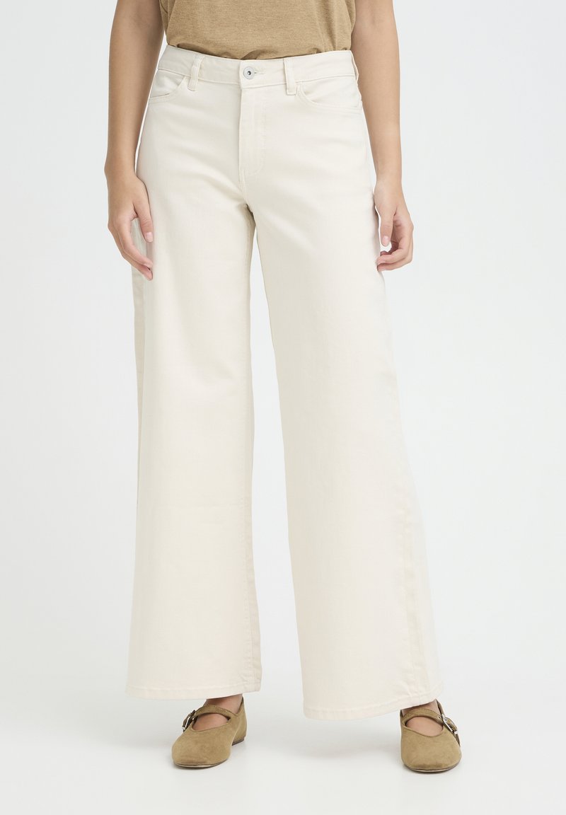 Pantalon beige à jambes larges avec fermeture boutonnée et à glissière, porté avec des ballerines Mary Jane en daim marron, sur une personne debout.