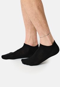 Schwarze Knöchelsocken aus dünnem Stoff, mit einem gerippten Bündchen, glatter Textur und minimalistischem Design, die auf baren Füßen getragen werden und sichtbares Beinhaar zeigen.