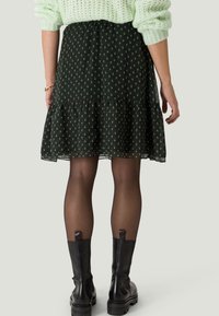 Jupe noire à motifs avec un ourlet volanté, présentant un design floral en vert. Portée avec des bottes en cuir noires à la cheville et des collants en voile.