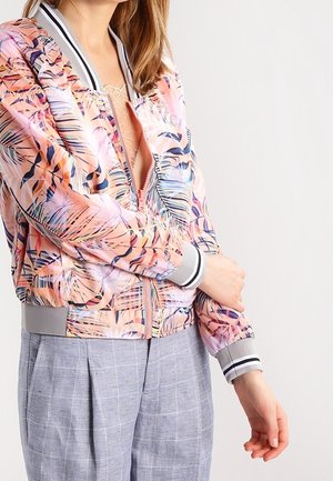 Vrouw die een bomberjack draagt in pastelroze en paarse patronen met grijze manchetten, gecombineerd met lichtgrijze geruite broek, terwijl ze haar mouw recht trekt.