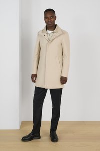 Abrigo de lana beige con cuello alto, bolsillos laterales y corte entallado, combinado con pantalones negros y zapatos, en un fondo minimalista.