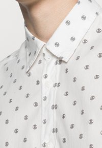 Camicia bianca con colletto a punta, caratterizzata da un motivo circolare nero su tutta la superficie. Il tessuto appare liscio con una vestibilità regolare.