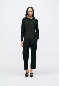 Crna majica s kapuljačom i dugim rukavima, uparena s crnim suženim hlačama. Model nosi crne cipele s oštrim vrhom. Učinkovita tekstura tkanine.