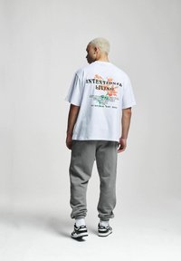 Tricou alb din bumbac cu inscripția "VIEȚI INTENȚIONAT" imprimată în negru și accente florale pe spate, asociat cu treninguri gri și adidași negri.