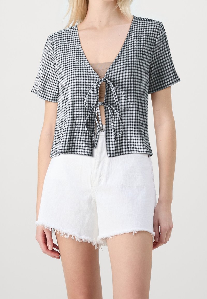 Top corto de cuadro vichy en blanco y negro con escote en V y lazo en el frente, mangas cortas, combinado con pantalones cortos de denim blanco desgastados.