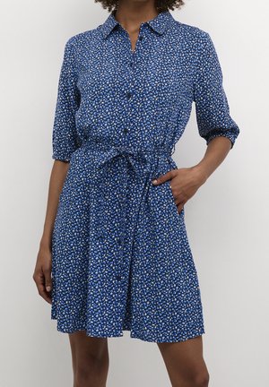 Kobieta ubrana w niebieską sukienkę koszulową z guzikami, w kwiatowy wzór, z przewiązanym paskiem w talii, jedna ręka w kieszeni, stojąca na jednolitym tle.