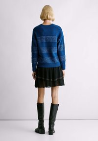 Suéter azul con rayas horizontales y acentos brillantes, combinado con una falda negra de capas y botas negras hasta la rodilla.