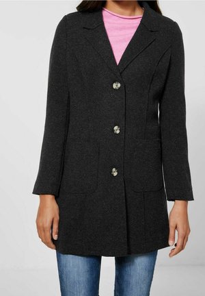 Femme portant un manteau boutonné gris foncé par-dessus un haut rose et un jean bleu, avec deux poches plaquées visibles à l'avant.