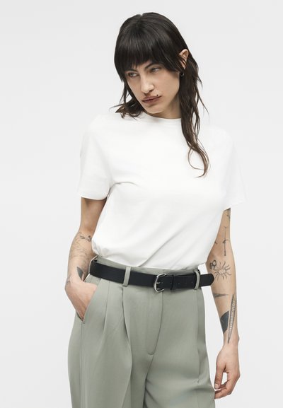 Άτομο με σκούρα μαλλιά που φοράει λευκό μπλουζάκι μέσα σε ψηλόμεσα γκρι παντελόνια με μαύρη ζώνη, ποζάροντας με το ένα χέρι στην τσέπη.