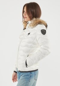Witte puffy jas met een met bont afgewerkte kap, rits aan de voorkant en twee zijzakken. Heeft een zwart logo-embleem op de linkermouw.