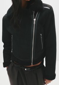 Giacca nera cropped con colletto in shearling, dotata di cerniere in argento, accenti in ecopelle e tessuto testurizzato. Include tasche laterali.