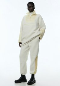 Cremefarbener Fleece-Pullover mit weichem, strukturiertem Finish und einem Reißverschlusskragen. Kombiniert mit passenden Hosen mit gelben Seitenelementen. Schwarze klobige Stiefel.