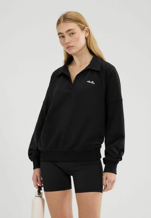 Kobieta w czarnym długim rękawie i czarnych szortach trzyma białą butelkę na wodę na jednolitym tle.